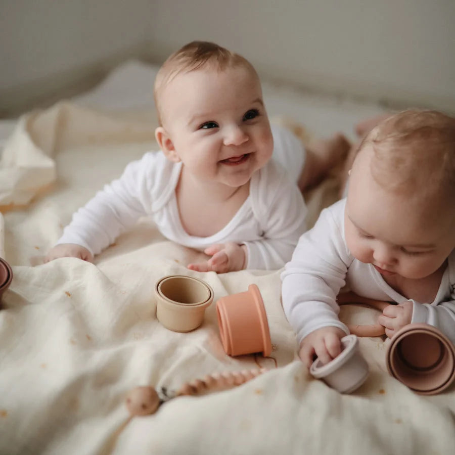 Zwei lächelnde Babys in weißen Overalls auf einem Bett mit Mulltuch Sun