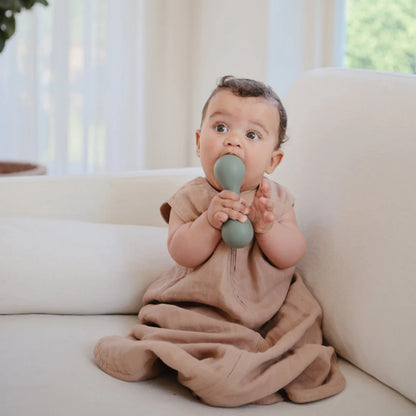 Baby sitzt auf einem Sofa und hält ein blaues Rasselspielzeug im Musselin Schlafsack