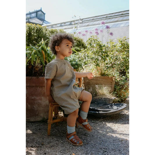 Kind trägt hellgraues kurzärmeliges leinenkleid mit knopfleiste und passende shorts zur cognacfarbenen oscar sandal