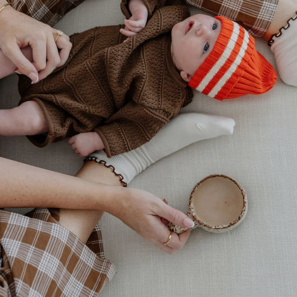 Baby in braunem Strickoverall und orange gestreifter Mützenöffnung