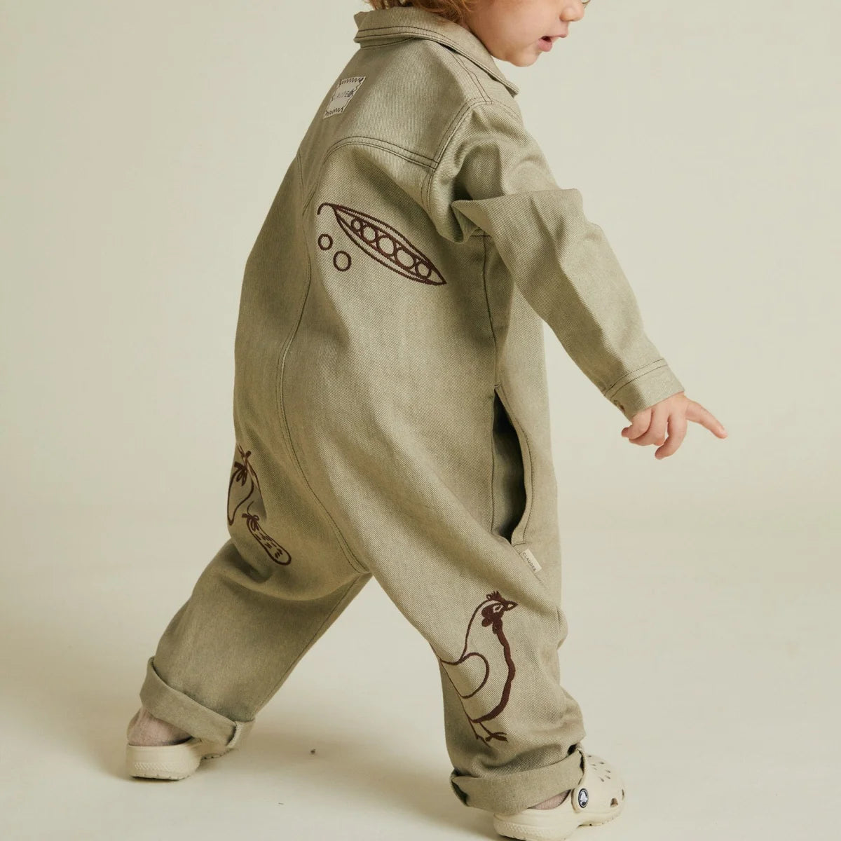 Ein kind trägt einen sagegrünen denim overall mit braunen stickereien von erbsen und hühnern quinn olive allotment