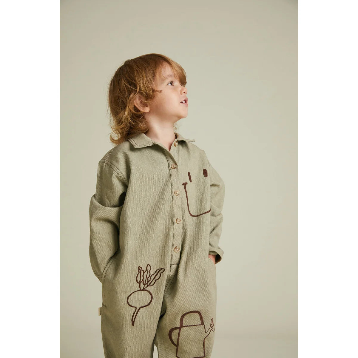 Ein kind trägt ein sagegrünes, langärmeliges overall mit braunen linienzeichnungen von gemüse und gartenwerkzeug