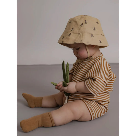 Baby in beige bucket hat with dark leaf patterns wearing copper stripes beach romper and matching socks holding green beans