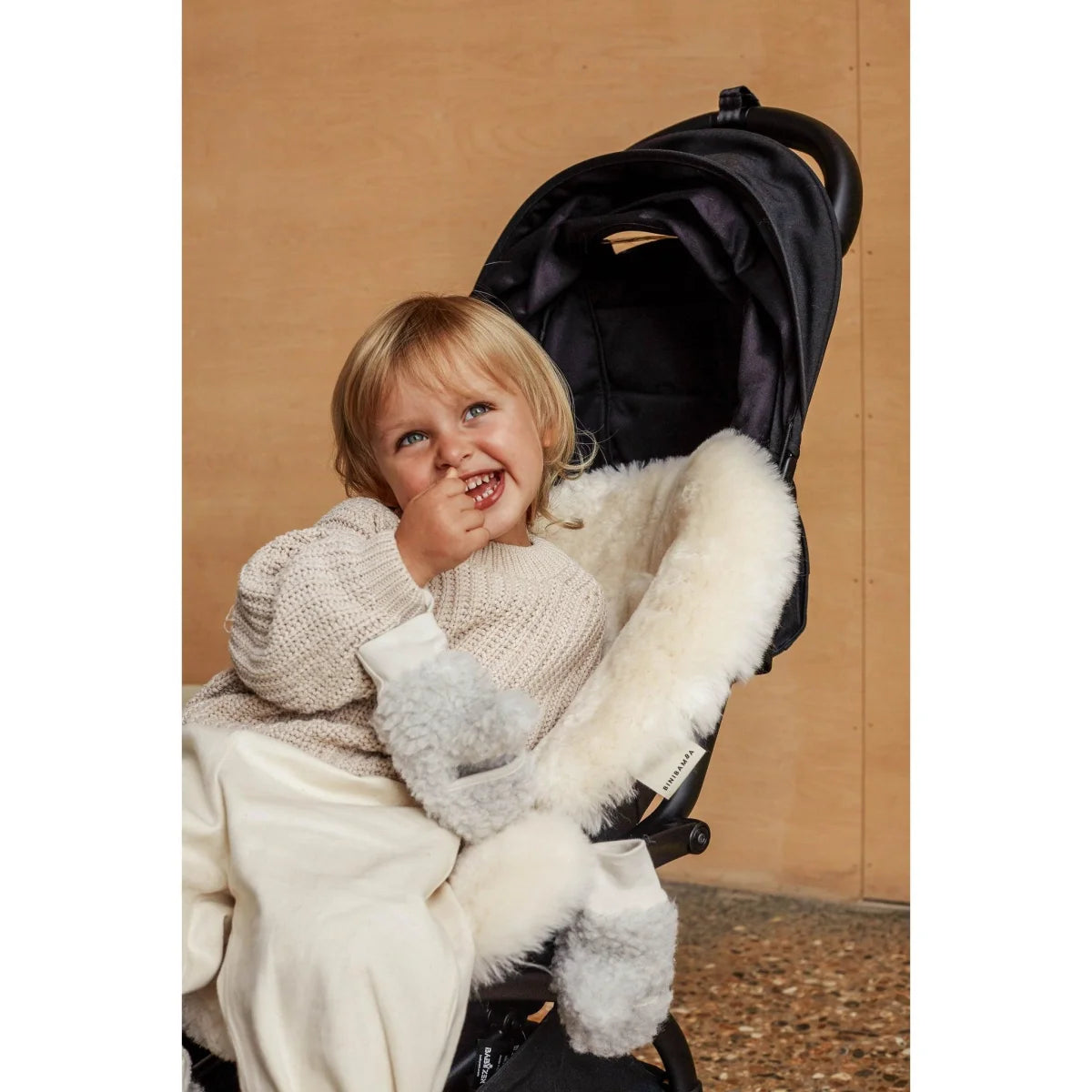 Kind im Kinderwagen, trägt einen weißen Pullover und lächelt mit Schaffell Snuggler