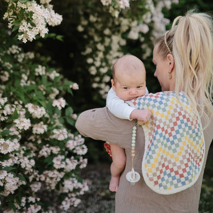 Baby mit Schnuller wird von einer Person in buntem Hemd bei blühenden Büschen gehalten