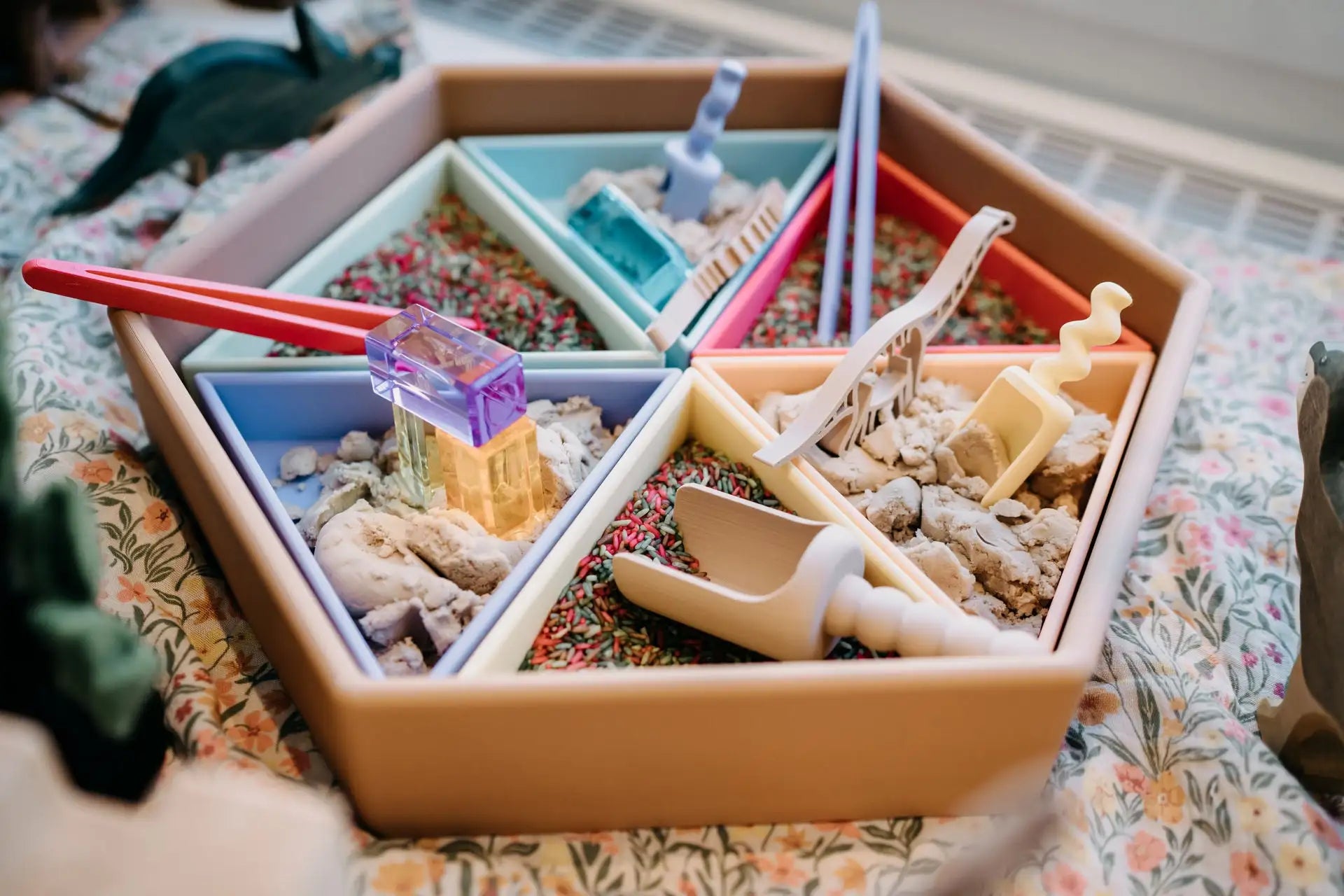 Sechseckige Sinnesbox mit farbenfrohen Fächern, in denen sich jeweils strukturierte Materialien und Plastikspielzeuge wie Schaufeln, Zangen und ein violetter, durchscheinender Block befinden.