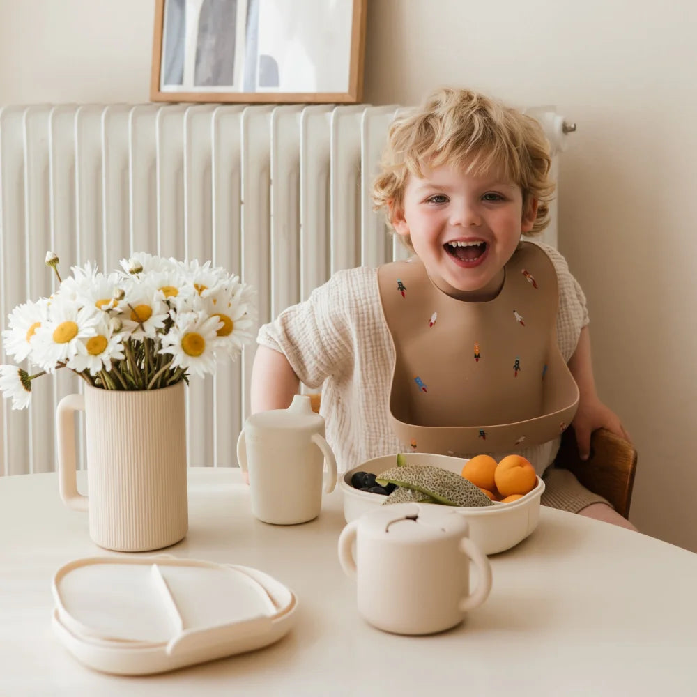 Lachendes Kind mit Lätzchen am Tisch mit Blumen und Essen, Produkt Snack Becher Ivory