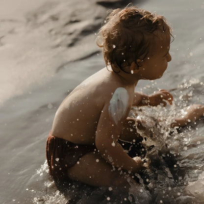 Kleinkind mit lockigem braunem Haar im flachen Wasser mit SPF 50+ Sonnencreme