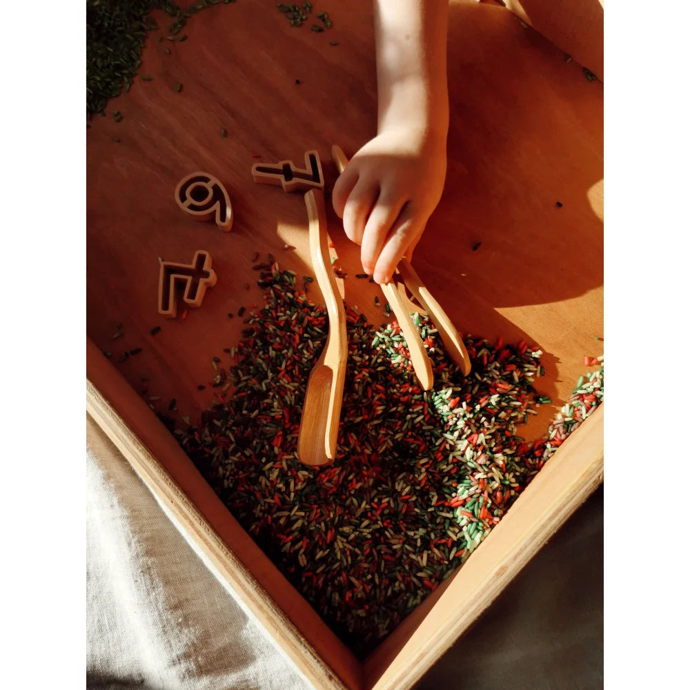 Kindliche Hand hält Holz-Sinnesspielzeug über buntem Reis und Holz-Zahlen in einer Spielwanne