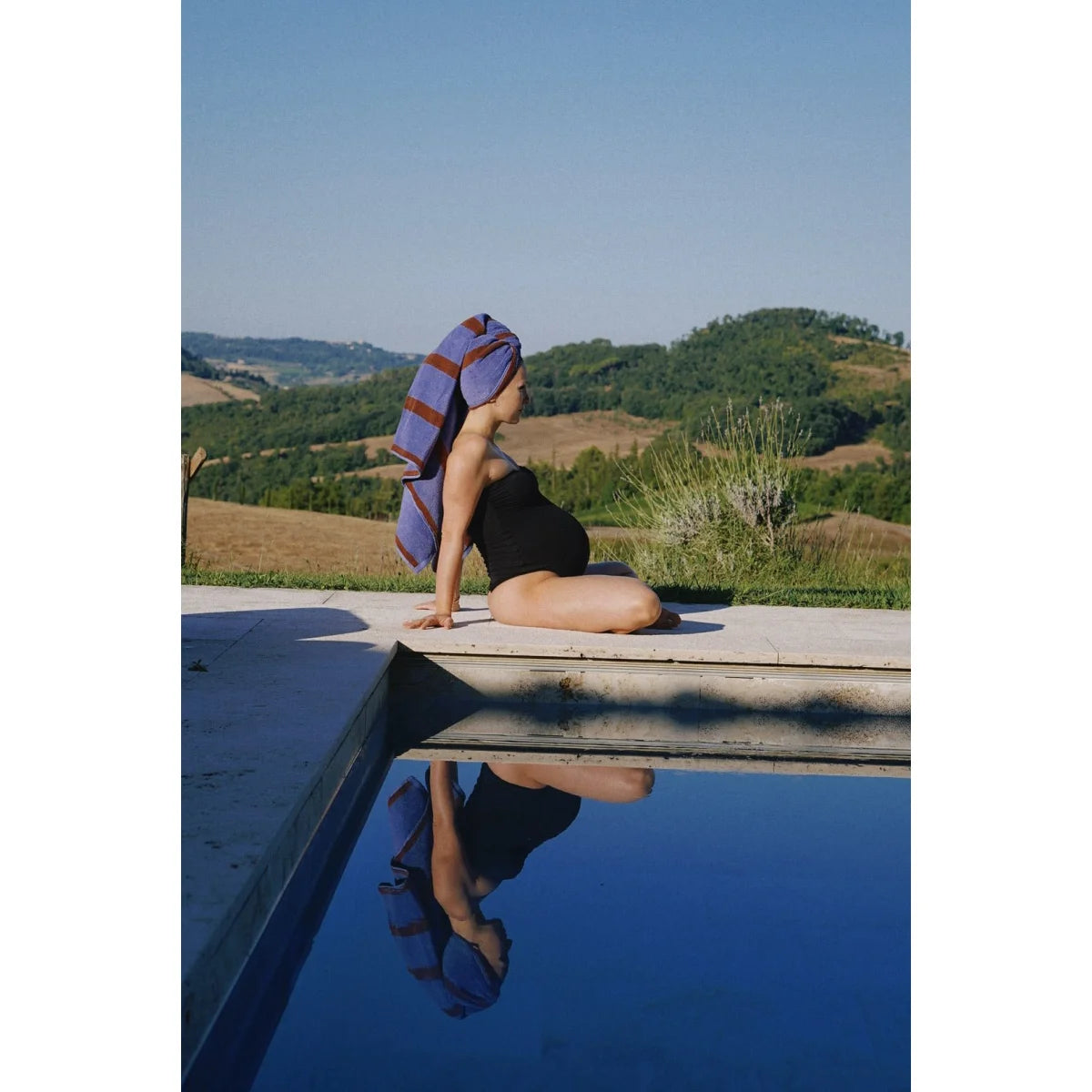 Eine schwangere frau sitzt am pool mit einem blauen und bordeaux gestreiften nachhaltigen badetuch als geschenk für neugeborene
