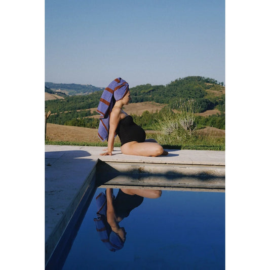 Eine schwangere frau sitzt am pool mit einem blauen und bordeaux gestreiften nachhaltigen badetuch als geschenk für neugeborene