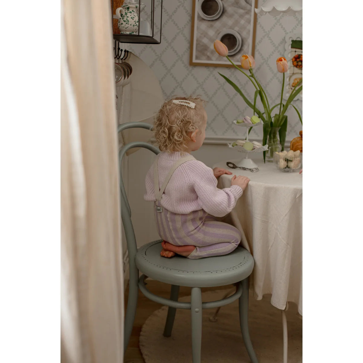 Ein kleines kind mit lockigem blondem haar trägt eine lavendel-creme strumpfhose mit trägern und sitzt auf einem grauen stuhl