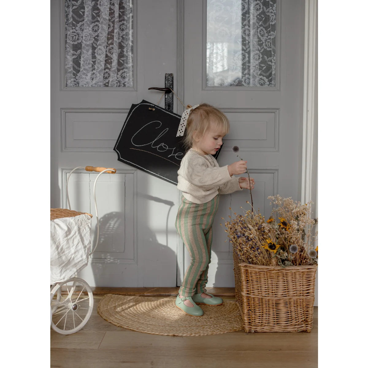 Ein kleinkind trägt strumpfhose mit träger footless in matcha und peanut neben einem korb mit getrockneten blumen