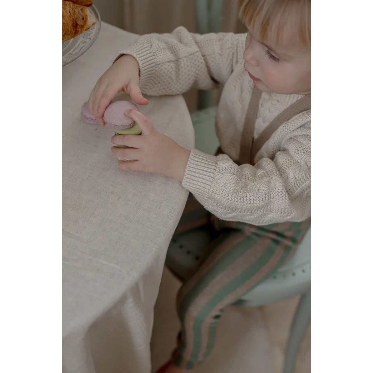 Ein kind hält zart ein rosafarbenes makron mit grüner füllung auf einem weißen tischtuch strumpfhose mit träger footless matcha x peanut