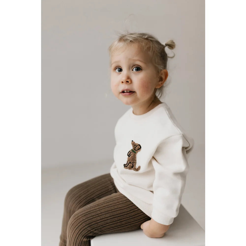 Kind trägt einen weißen Pullover mit Teddybär-Patch und braunen Hosen