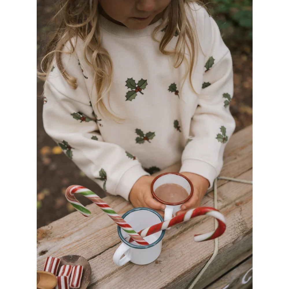 Kind trägt weißen Pullover mit Stechpalmenmotiven und hält eine Tasse mit Zuckerstangen