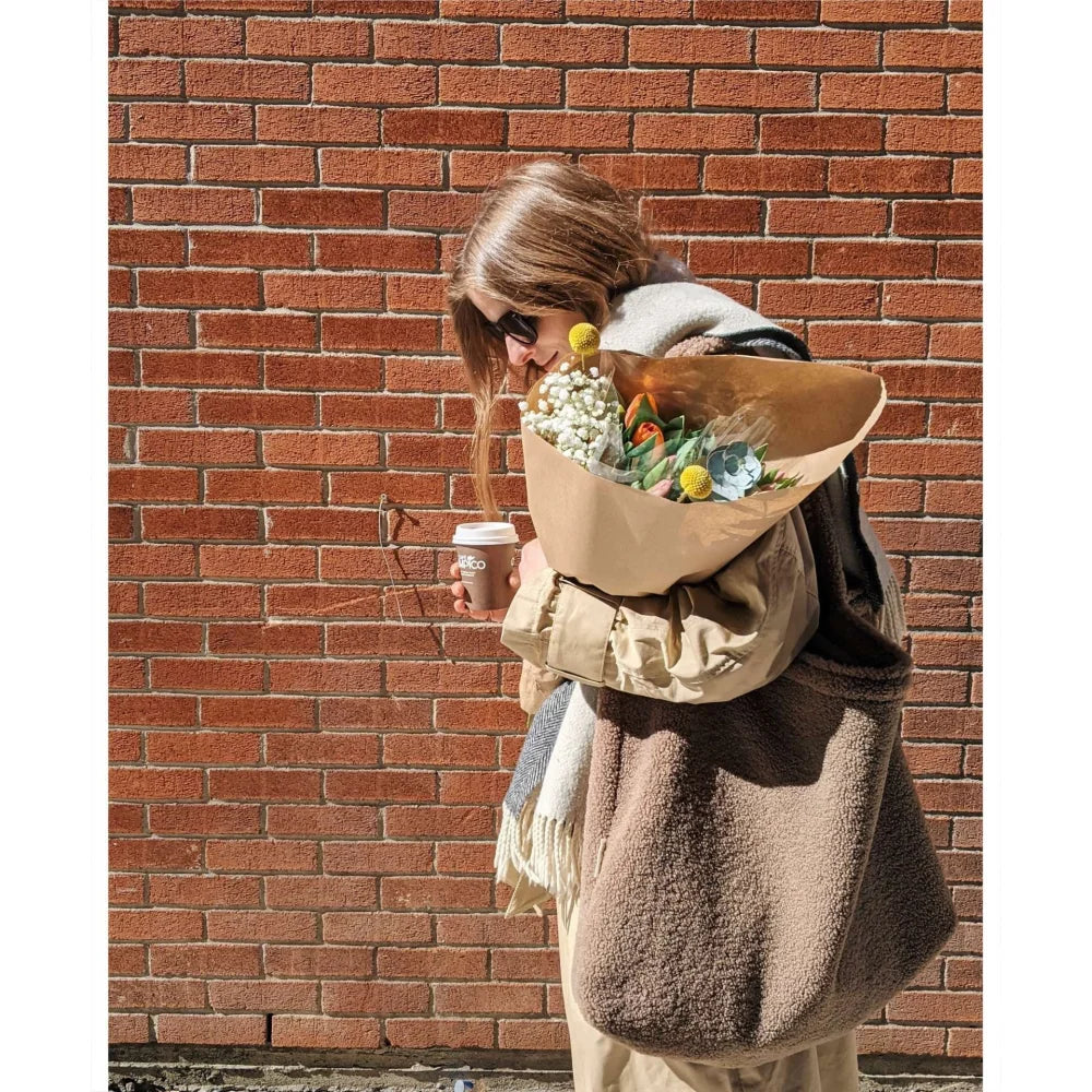 Frau mit Blumenstrauß und Kaffeebecher vor Ziegelwand mit Teddy Mom Bag in Braun