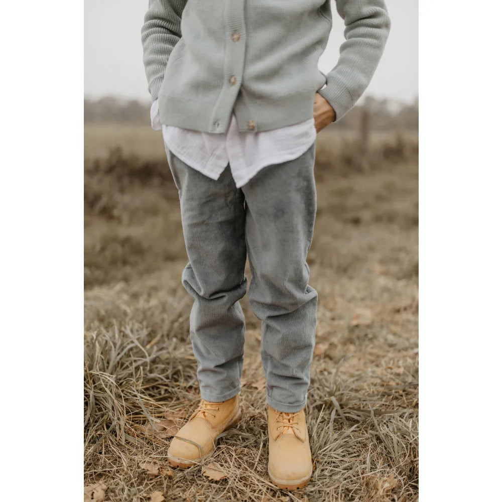 Junge trägt hellblauen Cardigan, weißen Hemd, graue Cordhose und braune Stiefel