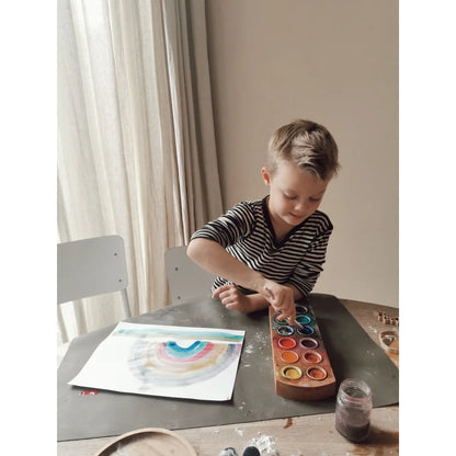 Junge mit hellen Haaren und gestreiftem Hemd malt Regenbogen mit Wasserfarben und Palette