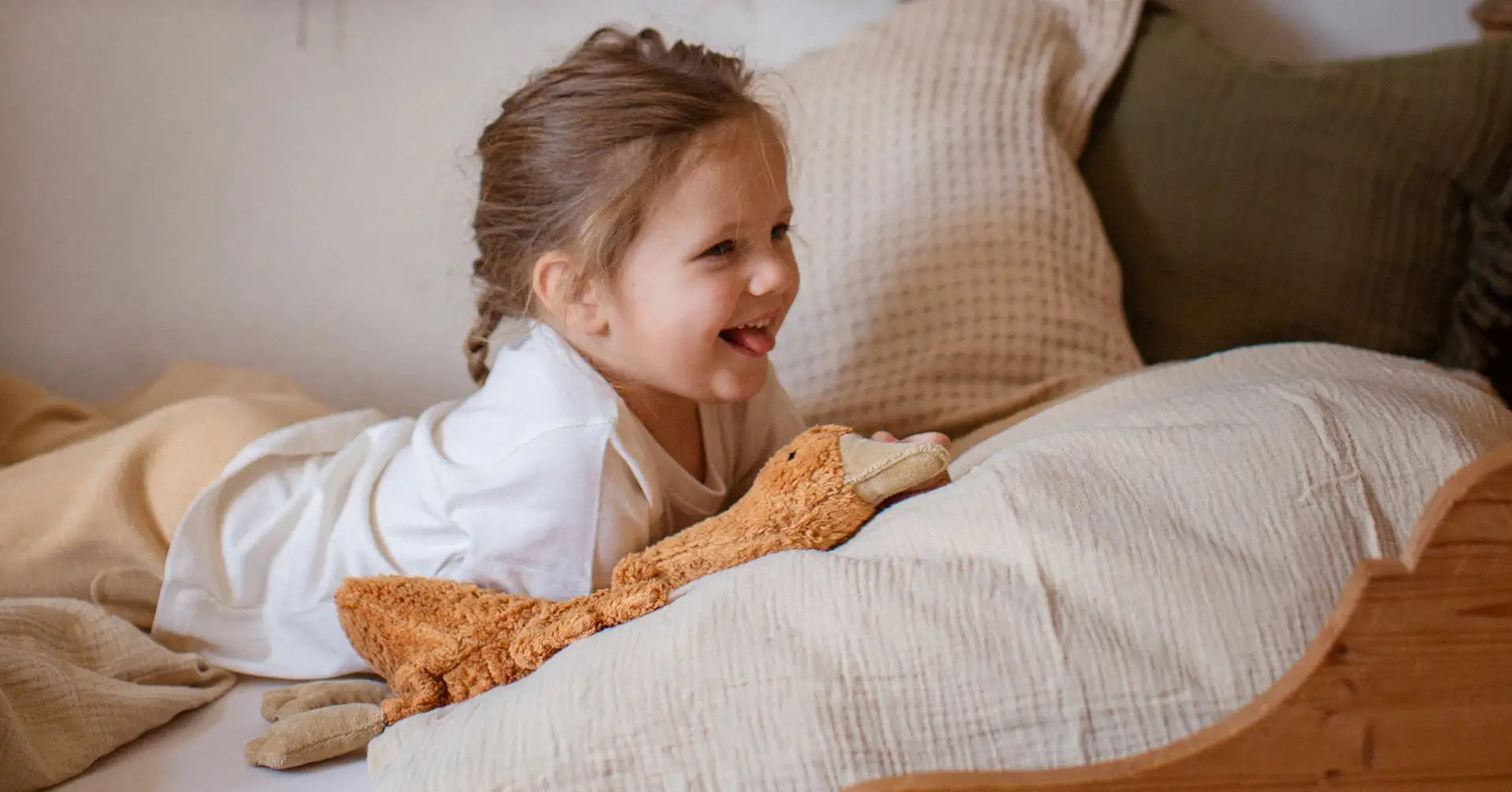 Ein weiches, hellbraunes Plüschgiraffen-Spielzeug mit einem texturierten Stoffkörper und hellfarbigen Füßen liegt auf dem Bett neben einem lächelnden Kind.