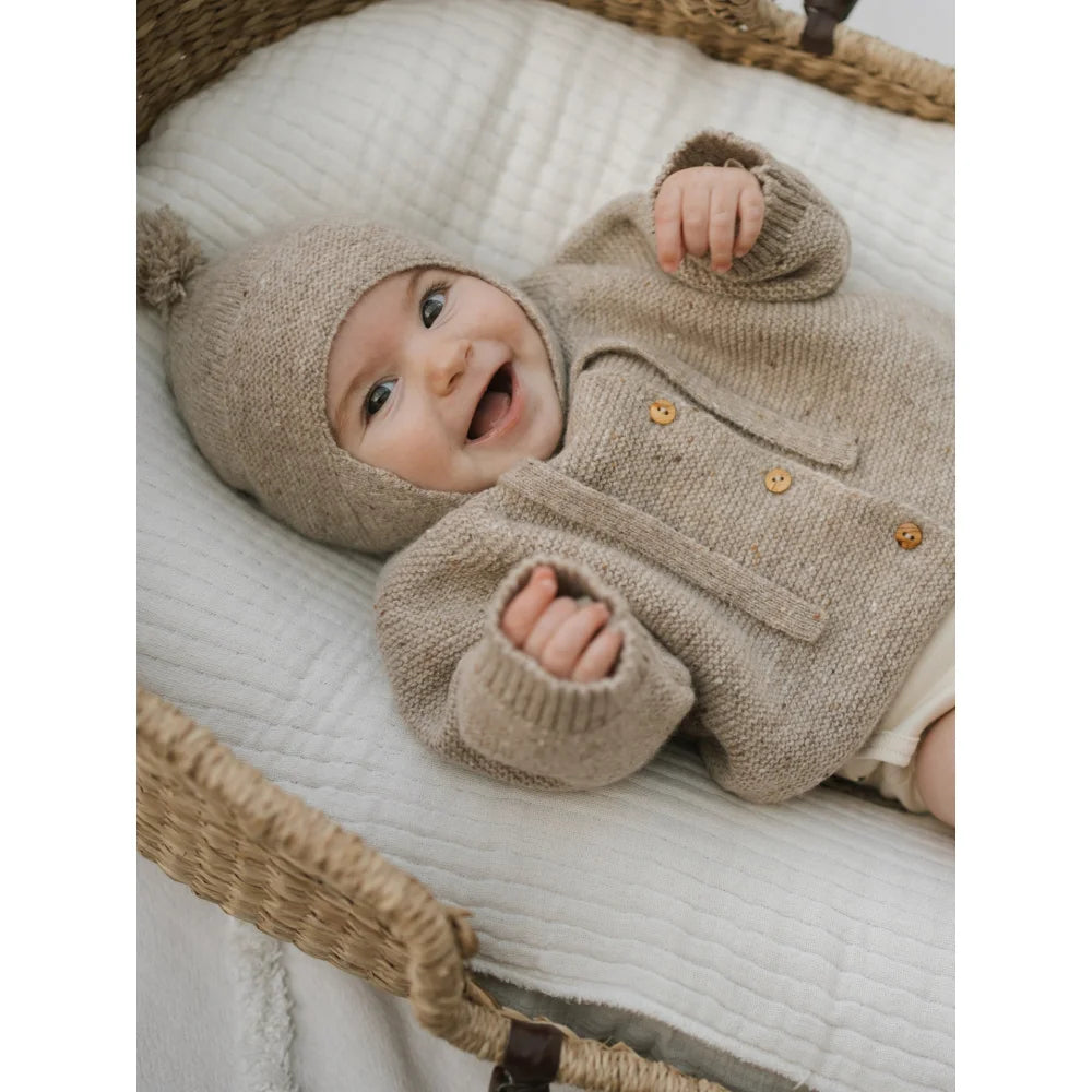 Lächelndes Baby in wolliger Oatmeal Strickjacke mit Pompon und Holzknöpfen
