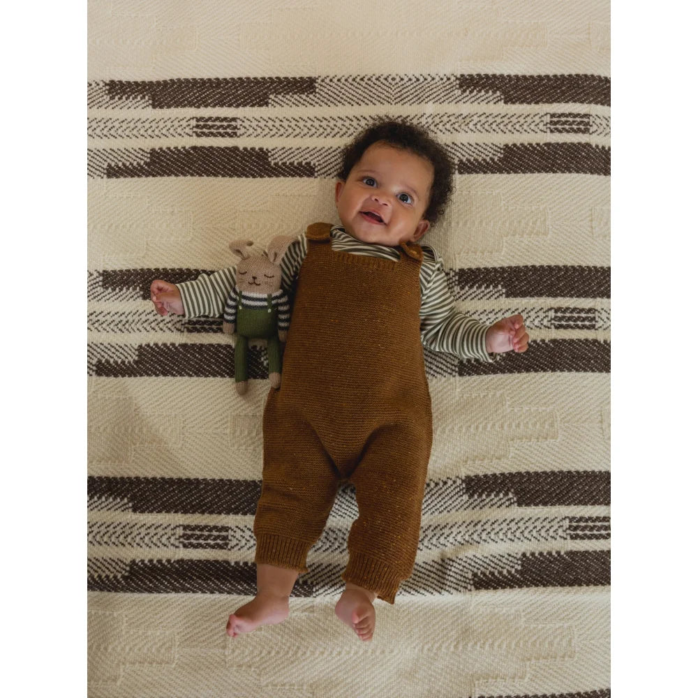 Kleiner Säugling mit dunklen Locken in Wool Salopette Chestnut neben Plüschhasen