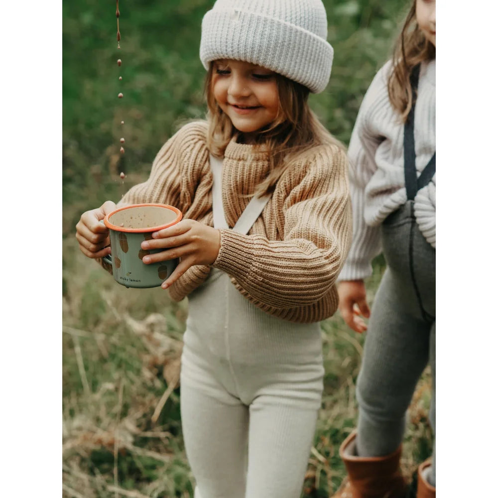 Junge mit weißer Mütze und beige Pullover hält Becher draußen, trägt Wollstrumpfhosen