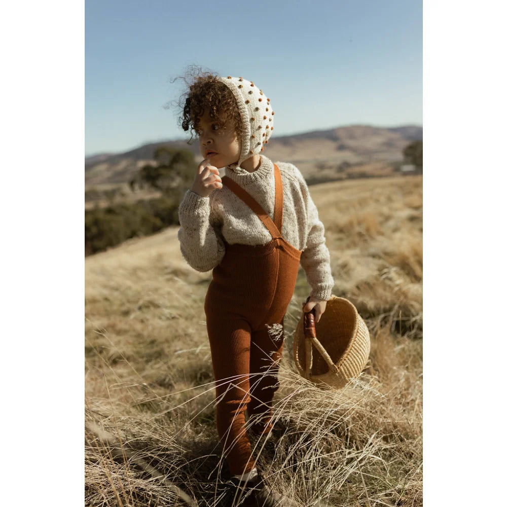 Kind trägt eine gestrickte Mütze und rustikale Overalls in einer Wiesenlandschaft